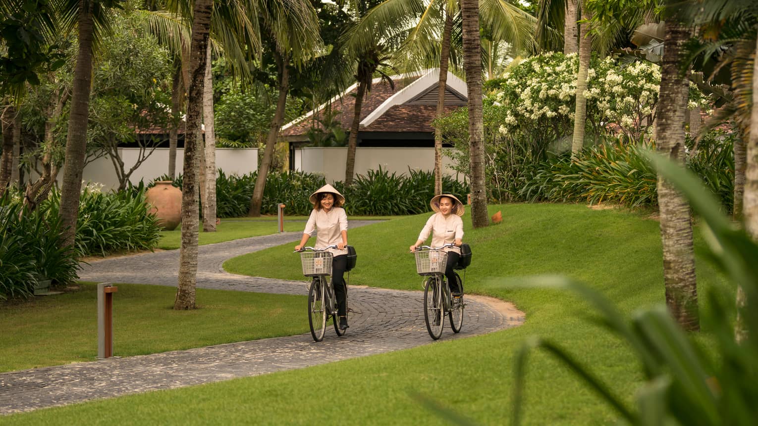 Two women wearing sun hats ride bicycles down winding brick path on resort 