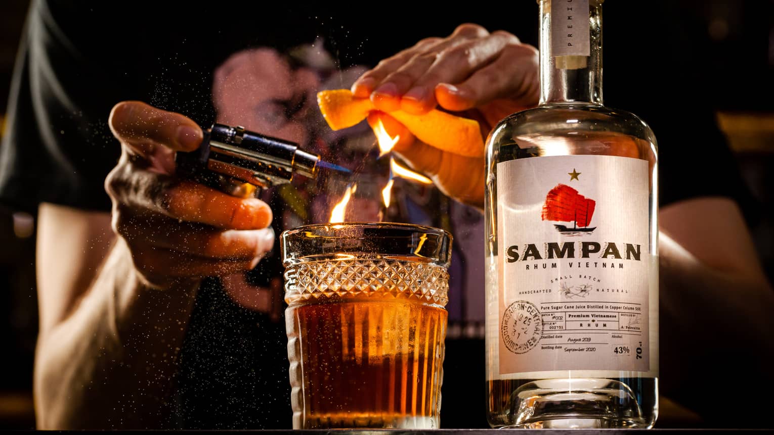 Person ignites an orange peel with a torch over a glass, bottle of Sampan rhum next to it