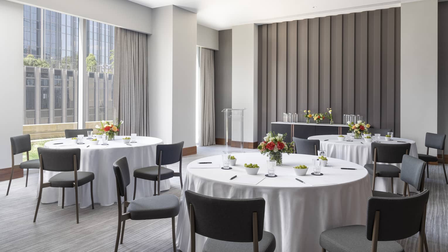 A meeting room with round tables, white tablecloths, dark grey chairs and a large window.
