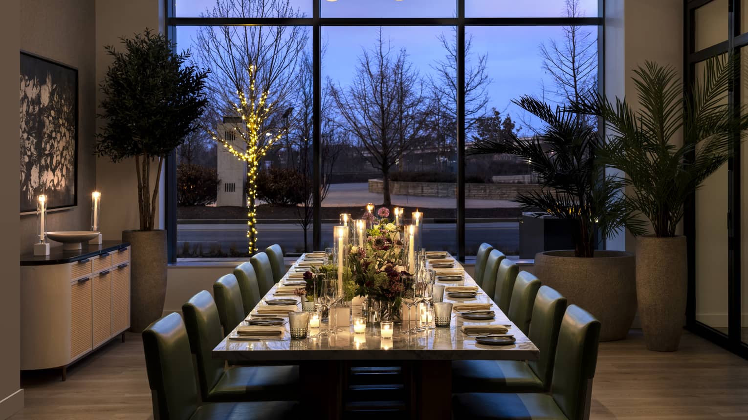 Dining table with candles and place settings, three large plants, and floor to ceiling windows.