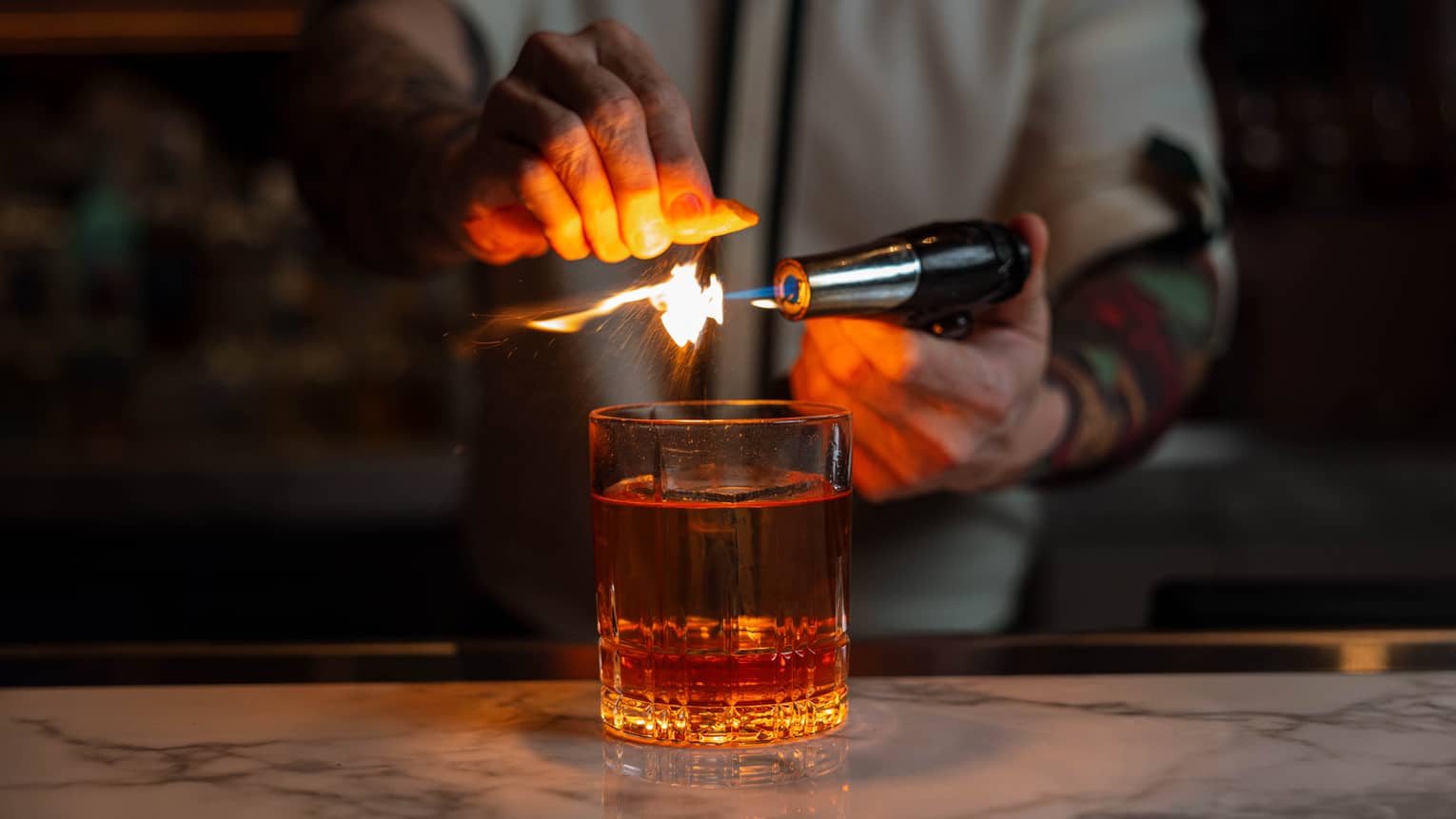 Bartender using a torch to flame an orange peel over a glass of whiskey, creating a dramatic effect at the bar.