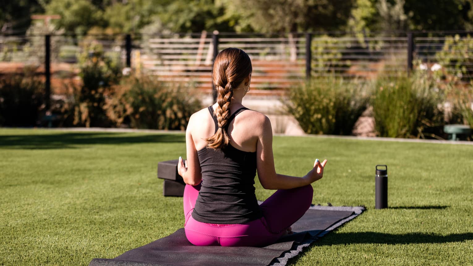 Woman wearing black tank top, pink yoga pants and her hair in a braid sits on a black yoga mat on a green lawn with her back to the camera