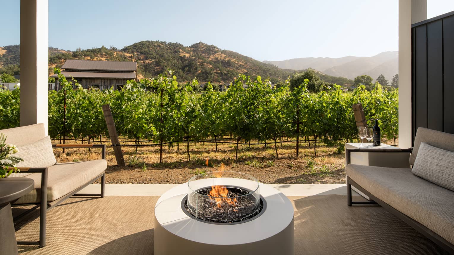 Covered patio featuring a gas fireplace in between two sofas, set next to lush vineyards