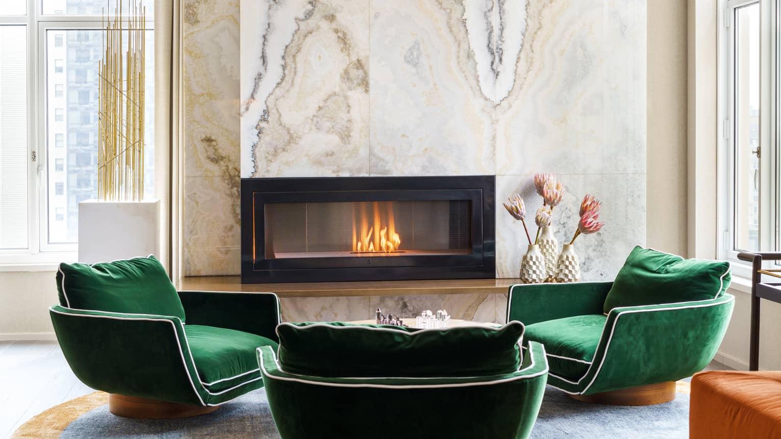 Three emerald-green velvet arm chairs by a fireplace, with marble accent wall