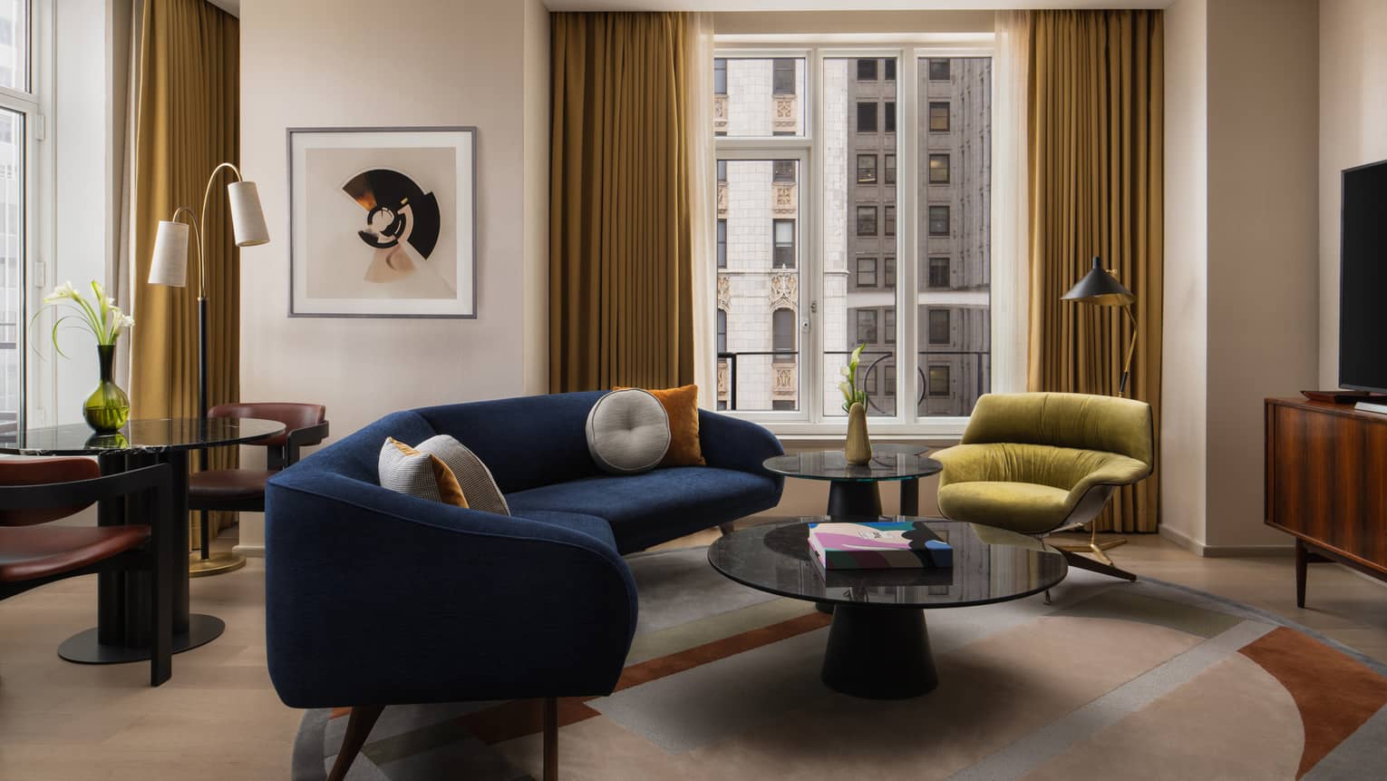 Living room with a navy blue sofa, green lounge chair, round coffee tables, abstract artwork and floor-to-ceiling windows with gold curtains