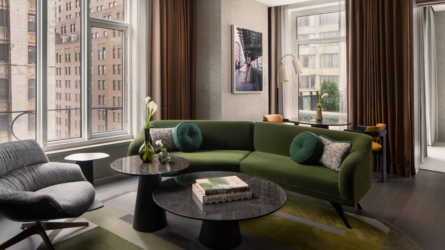 Luxury hotel suite living room with a green velvet sofa, round coffee tables, armchair, wall art and large windows with views of city buildings