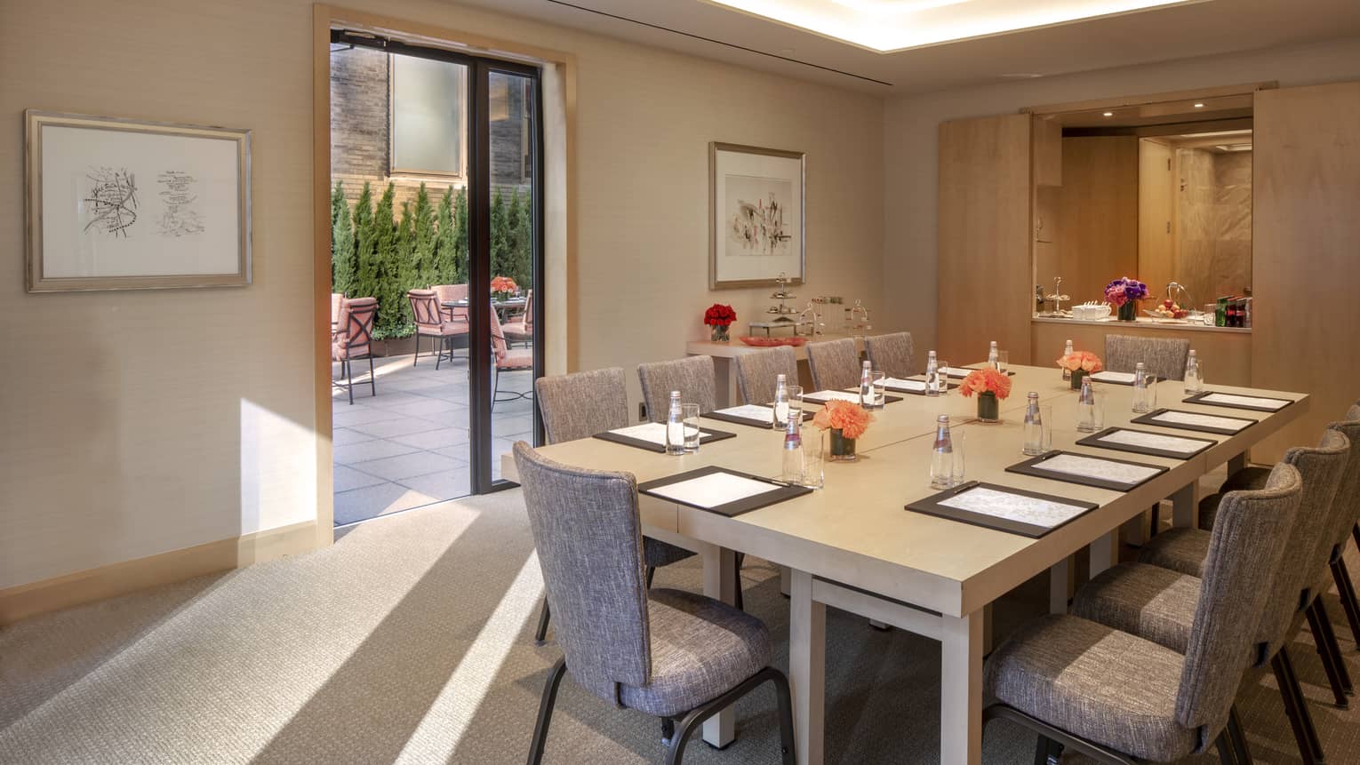 a tan rectangular table is set with an empty water glass, black folder, paper and pen. The door to the outdoor terrace is open and the light is pouring in