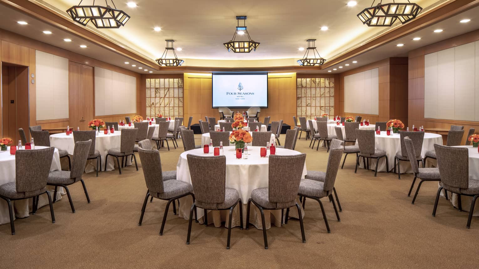 A large, wood panelled function room with circular tables set with a white tablecloth, red cups and orange flowers