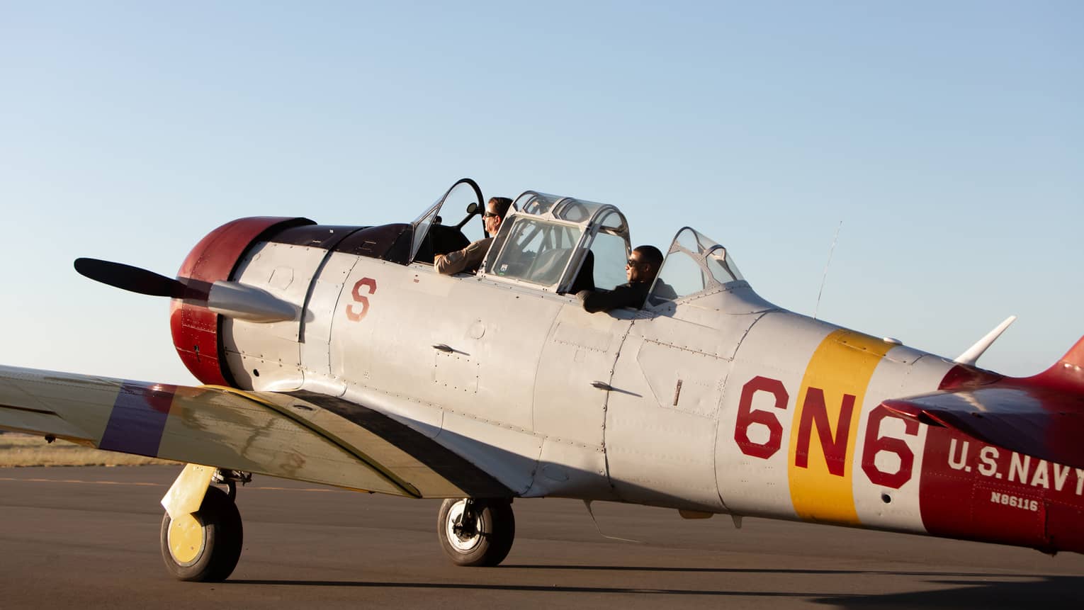 Two people sit in the cockpit of a vintage U.S. Navy aircraft on a runway, preparing for takeoff under a clear sky.