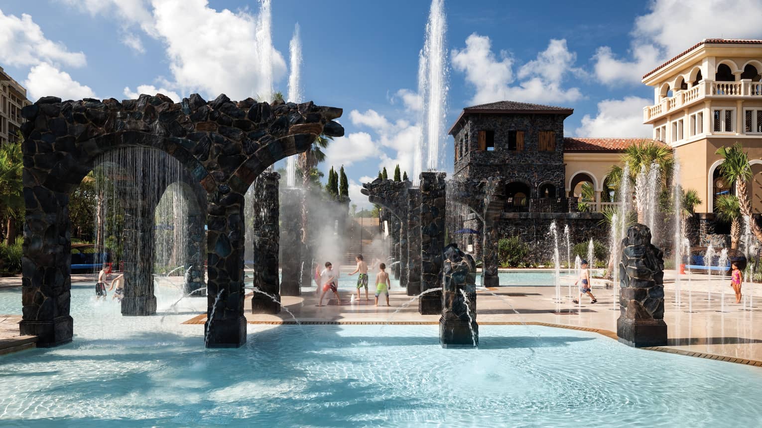 Kids play in water, under sprinklers and stone pillars in the Splash Zone water park