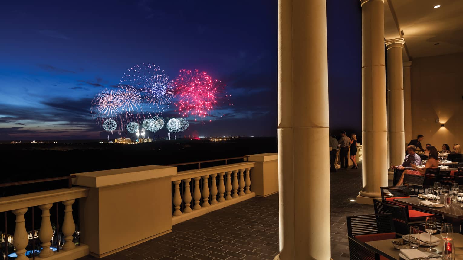 The terrace of a restaurant with tables and people watching fireworks in the distance.