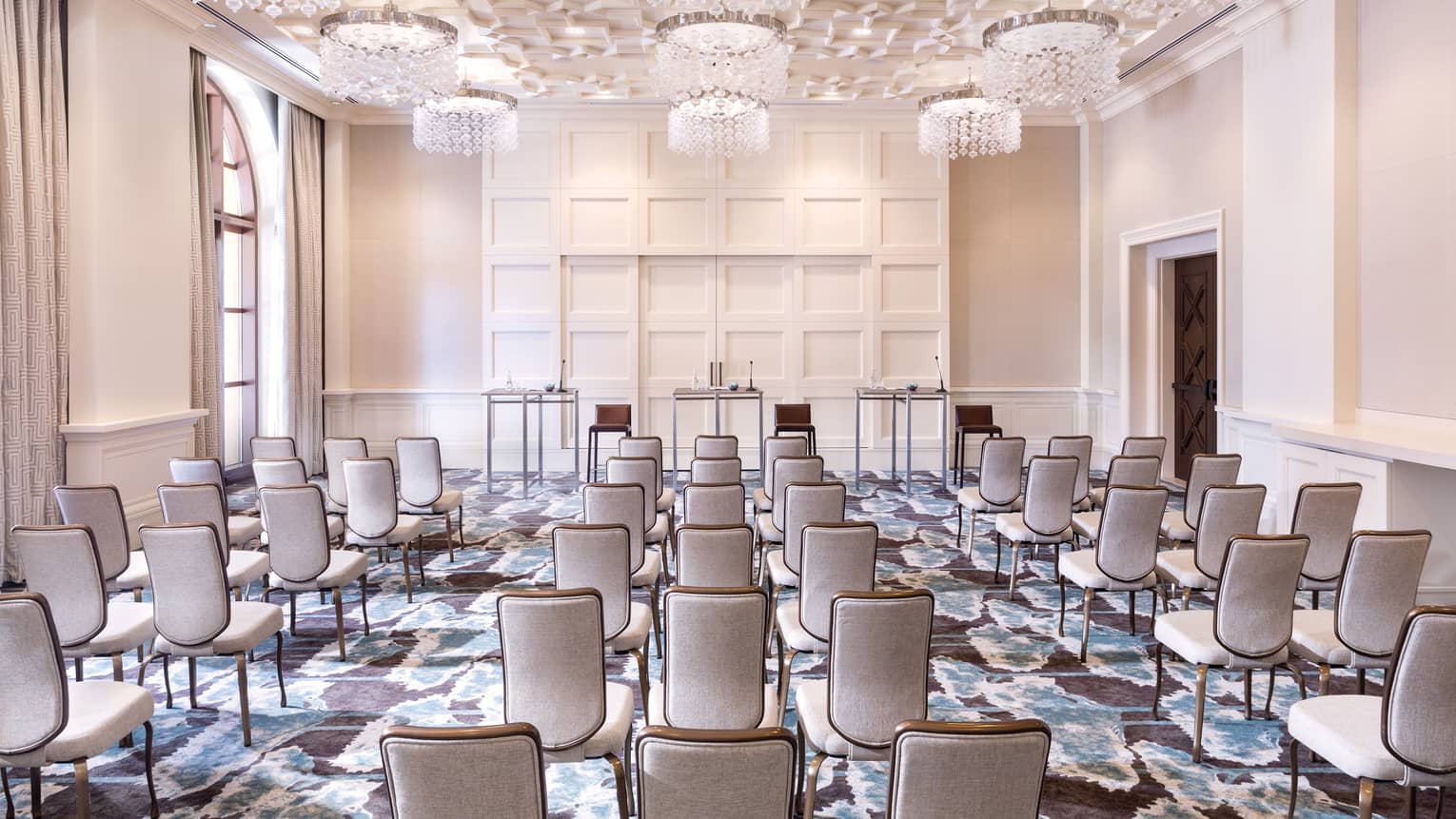 A cream colored function room is set up with rows of neutral chairs lined up and facing three tables in the front