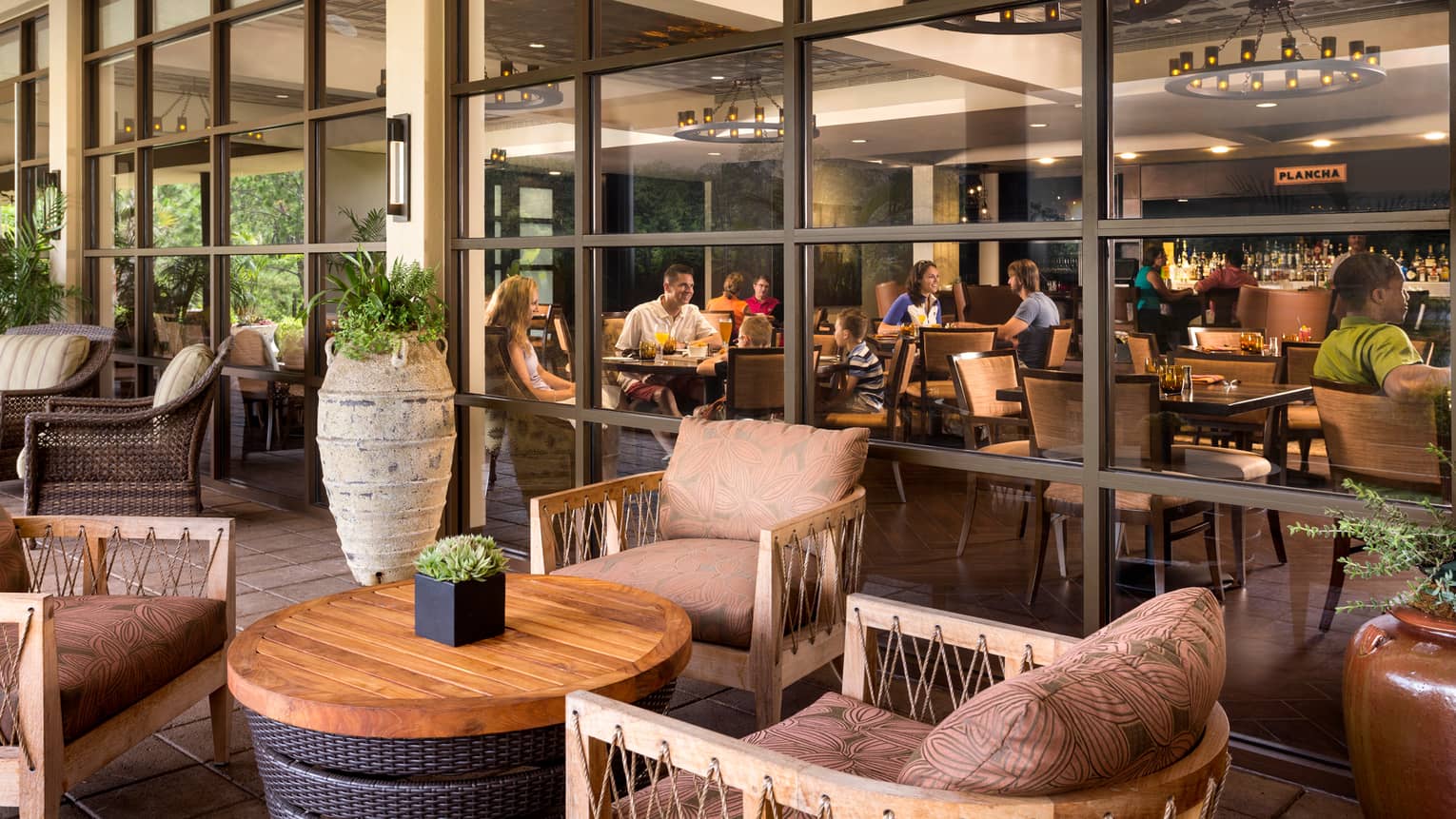 Plush patio chairs around wood table, tall vase, glass wall showing diners at tables