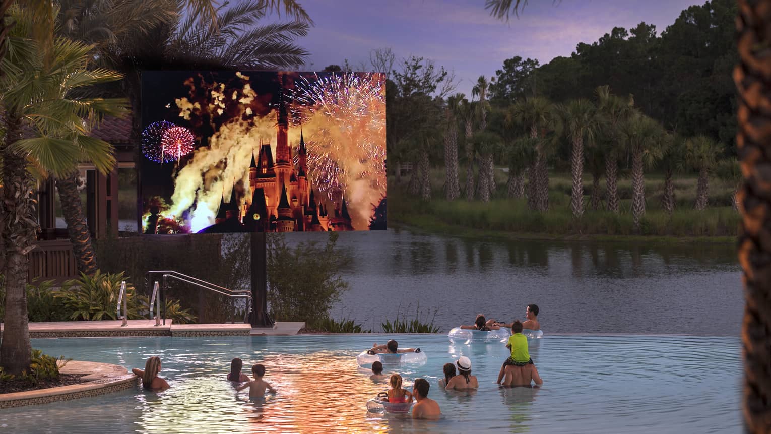 Guests watch a large screen displaying fireworks exploding over Cinderella's castle while floating in an outdoor pool