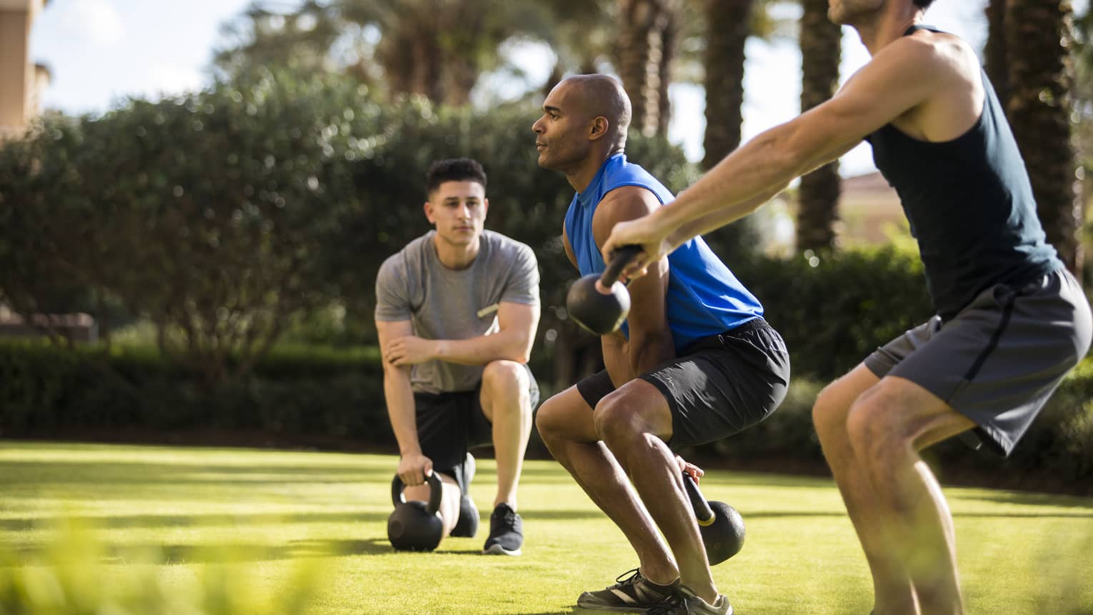 Four Seasons trainer watches as two men squat, lift kettlebell weights on lawn