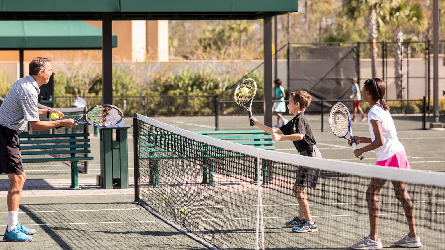 Man serves tennis ball to two kids on other side of net on small tennis court