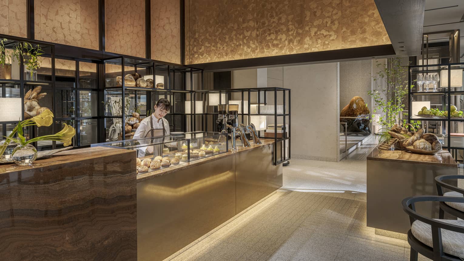 A person stands behind a counter displaying food items in an open, well-lit space decorated in warm neutral tones; the space also includes chairs, open shelves, an additional counter and a freestanding island