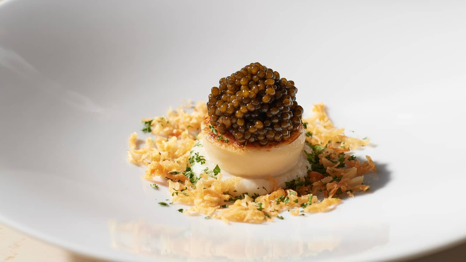 Round white plate with single scallop topped with caviar, surrounded by breadcrumbs and a smattering of green herbs