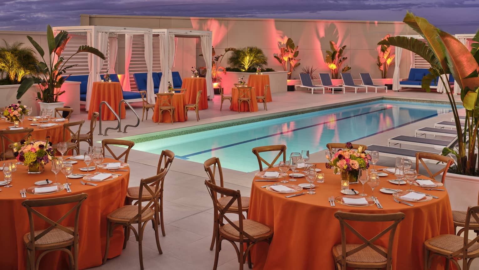 Pool with tables and chairs that have orange tablecloths and cabanas.