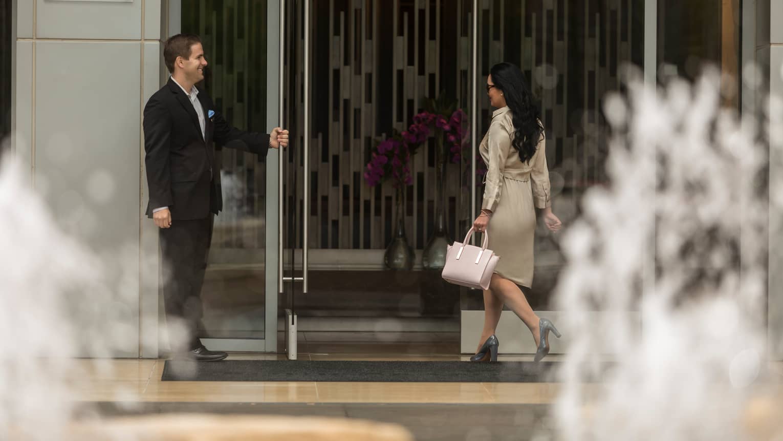 Man wearing suits holds glass door open for woman wearing designer dress, purse