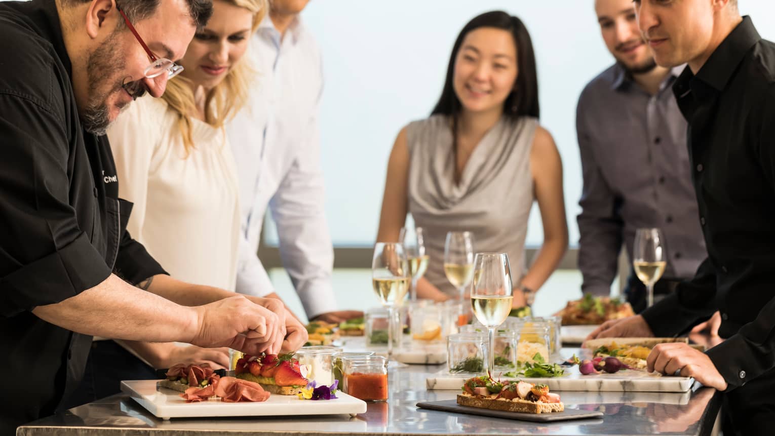 Chef assembles food at counter with smiling group of friends, white wine in glasses