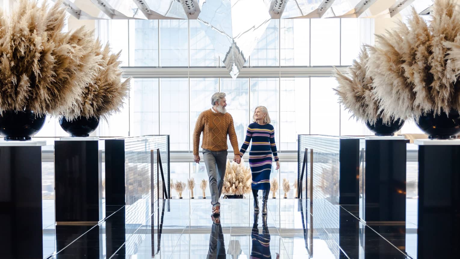A man and woman holding hands while walking in a large room covered in glass windows.