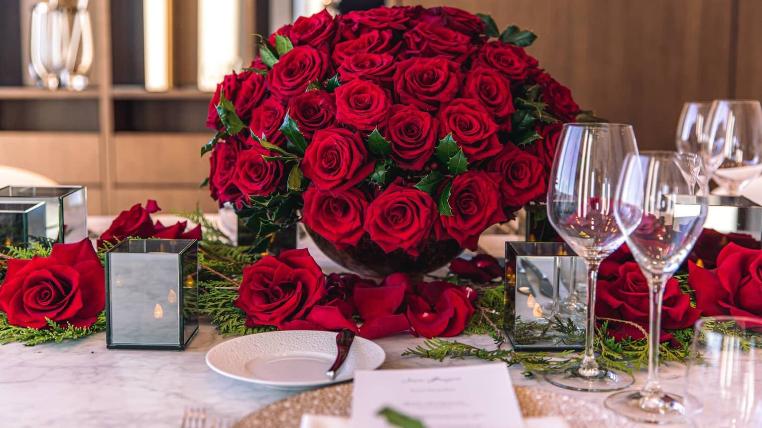 Red roses covered the edge of a table with white plates and wine glasses on it.