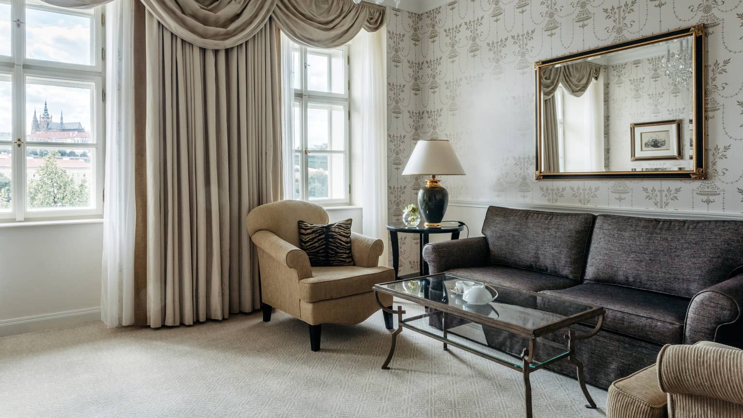 Room with brown leather sofa and mirror behind on wallpapered wall, two tan chairs, chandelier and curtains
