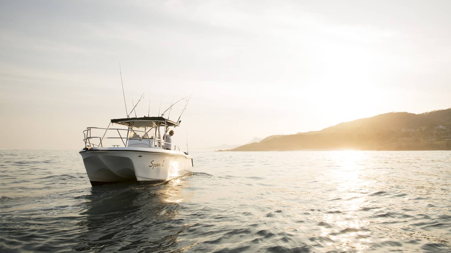 Front long view of a white boat bobbing in gentle waves with a person onboard looking out at the sun reflecting on the water.