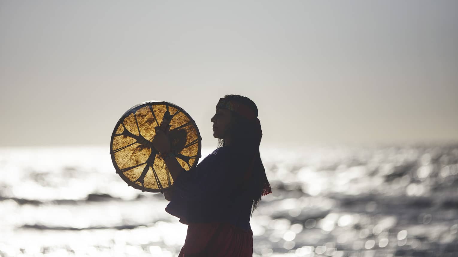 A woman holding a woven disc by the ocean.