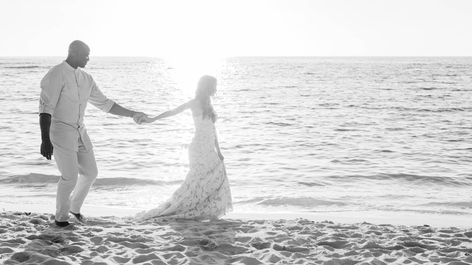 A bride and groom walk hand in hand down a beach at sunset