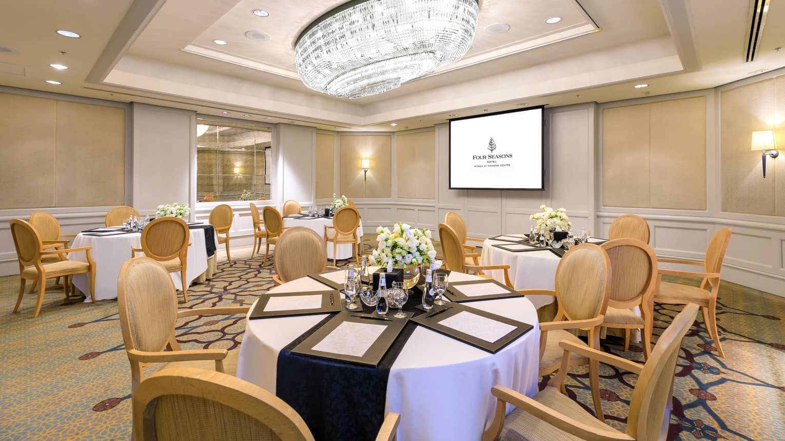 Elegant hotel meeting room with six banquet tables, presentation screen and crystal chandelier
