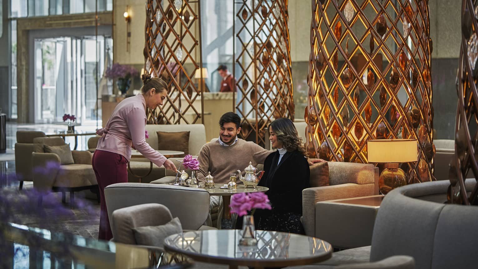 Two people sit a table in the middle of a lobby lounge while a server pours them tea