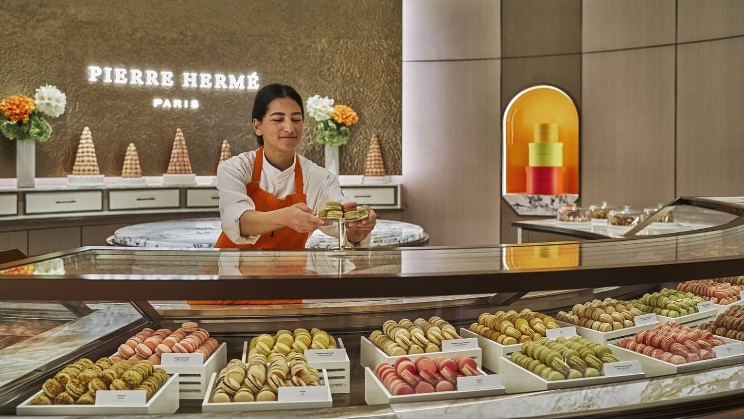 Bakery attendant stands behind a curved glass display case filled with macarons