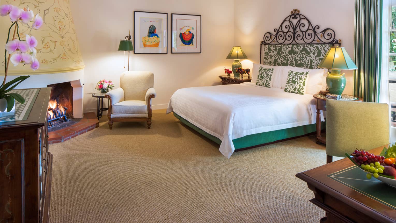 Jungle Room bed with iron and green-padded headboard, white armchair by fireplace, desk, fruit bowl