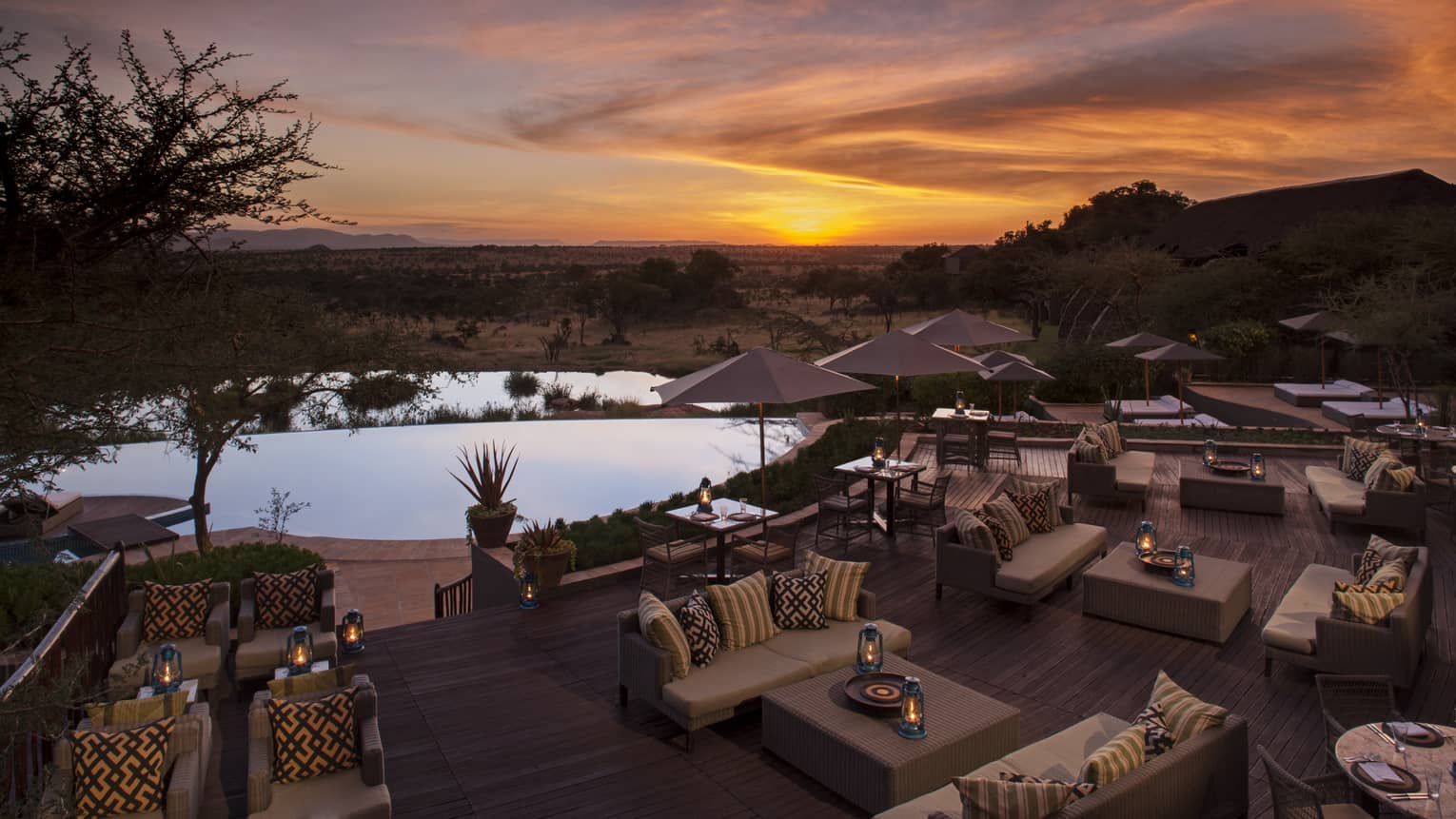 Outdoor furniture with pillows surround low tables, hurricane lanterns on patio overlooking infinity pool at dusk