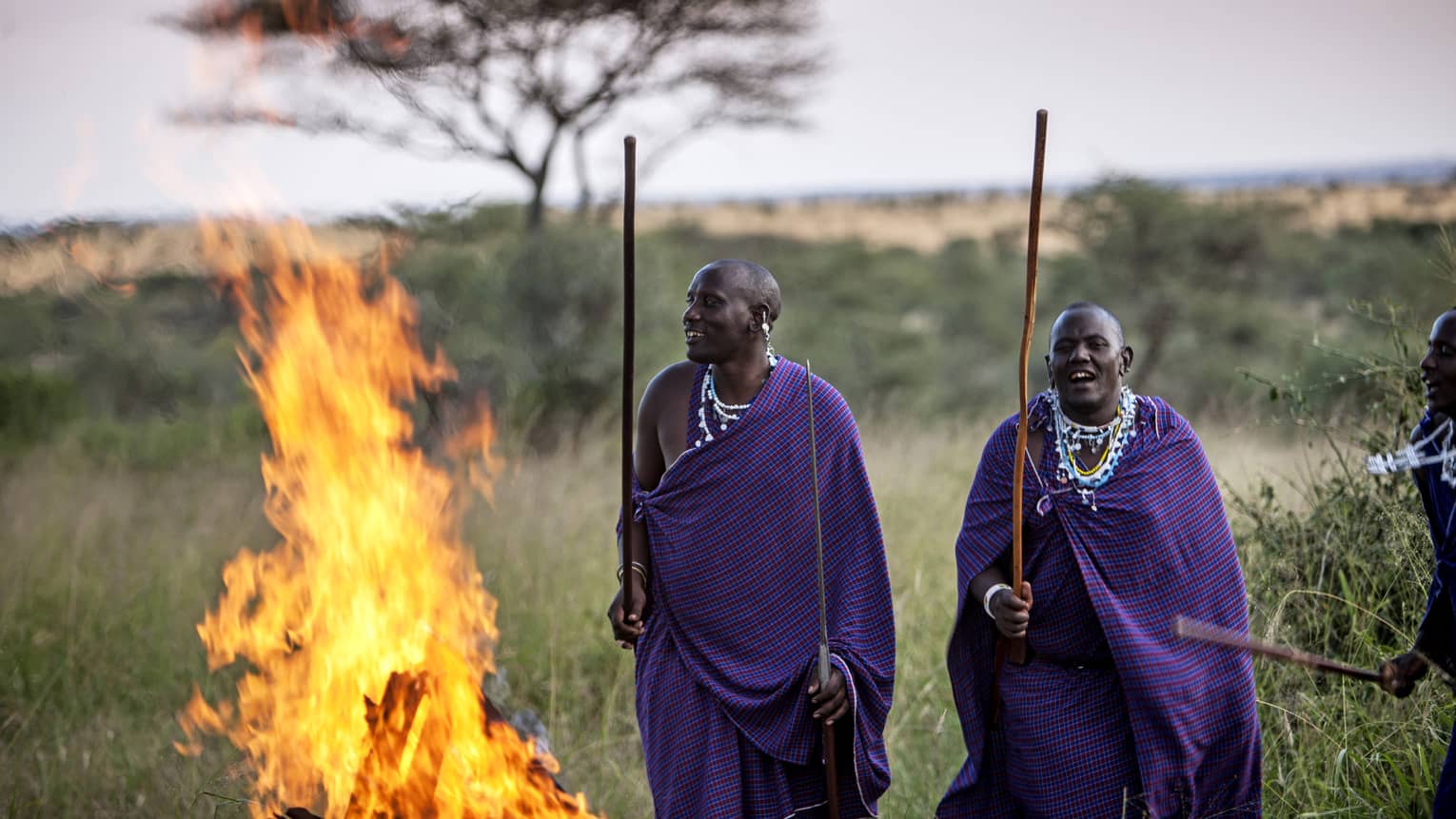 Three Maasai men holding sticks stand beside fire in the Serengeti 