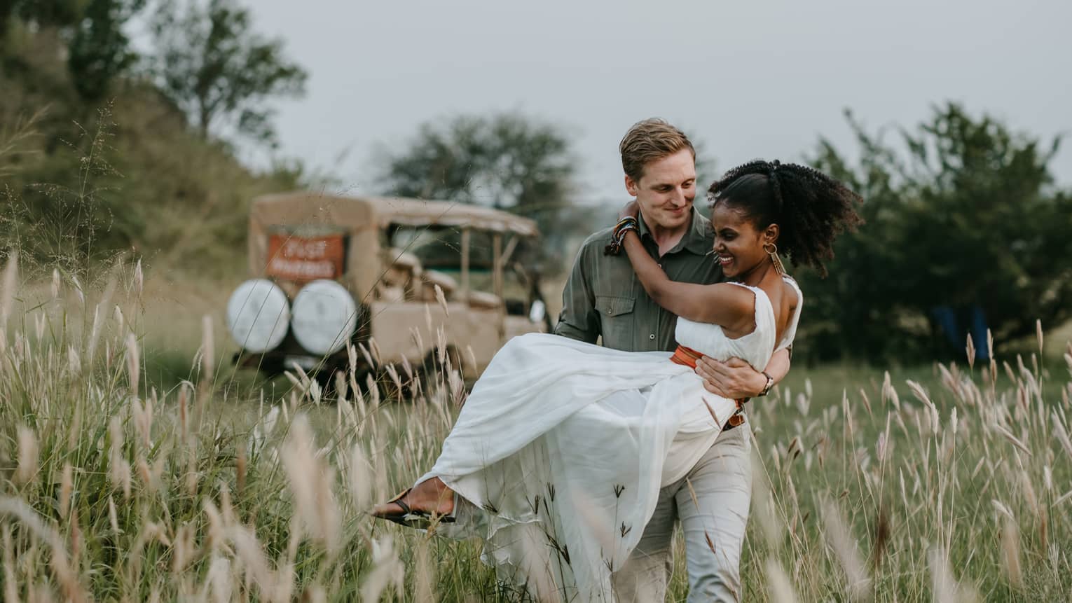 Man wearing olive green shirt and khaki pants holding woman in long white dress with red sash in his arms in a bush picnic setting