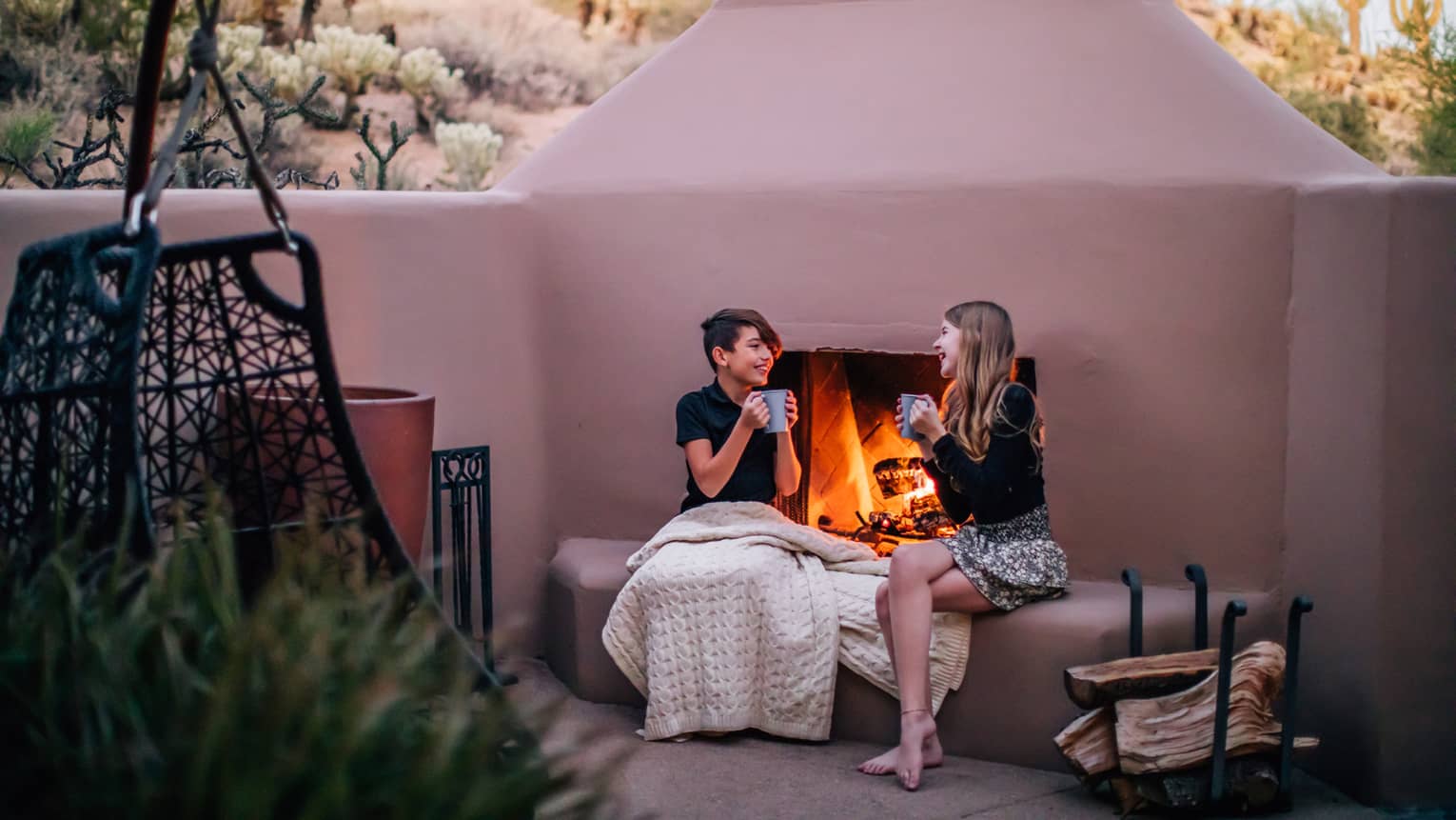 Two people sitting by a fire outside while drinking from mugs.