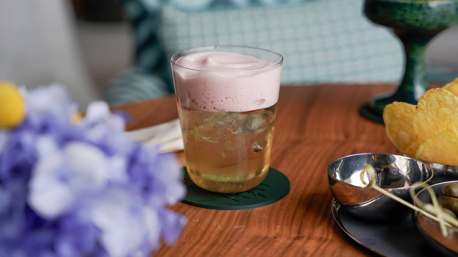 A cocktail topped with a light-pink foam sits in the centre of a wooden table next to purple flowers and a platter of appetizers
