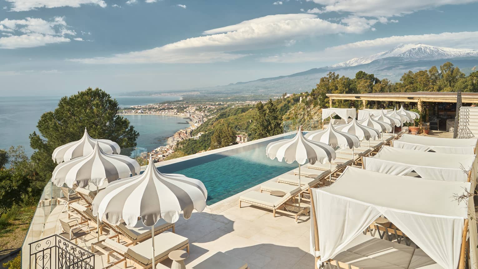 Infinity pool overlooking the sea shore surrounded by grey and white pinstripe pool umbrellas and loungers.