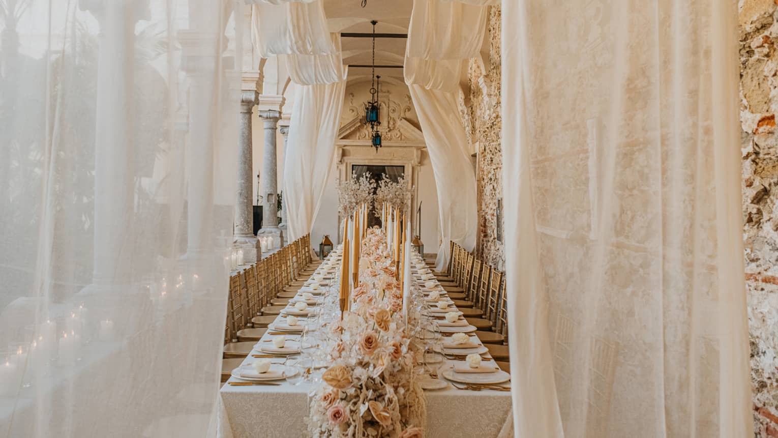 A long spacious event space with long tables set for a celebration, including peach and white floral arrangements all down the table's centre and peach and white taper candles in candlesticks. Gauzy white draping accents the scene.