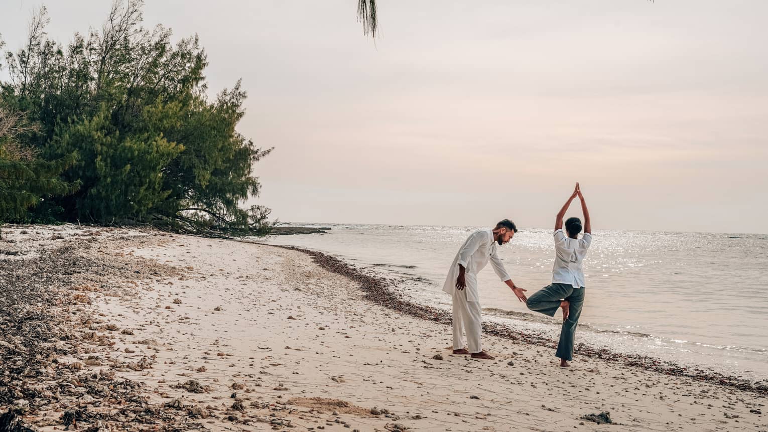 Yoga instructor assists student doing tree pose on the beach