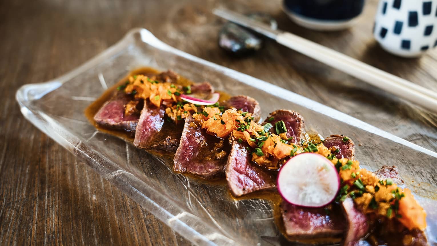 A dish of Waygu Tataki served on a clear plate with chopsticks in the background.
