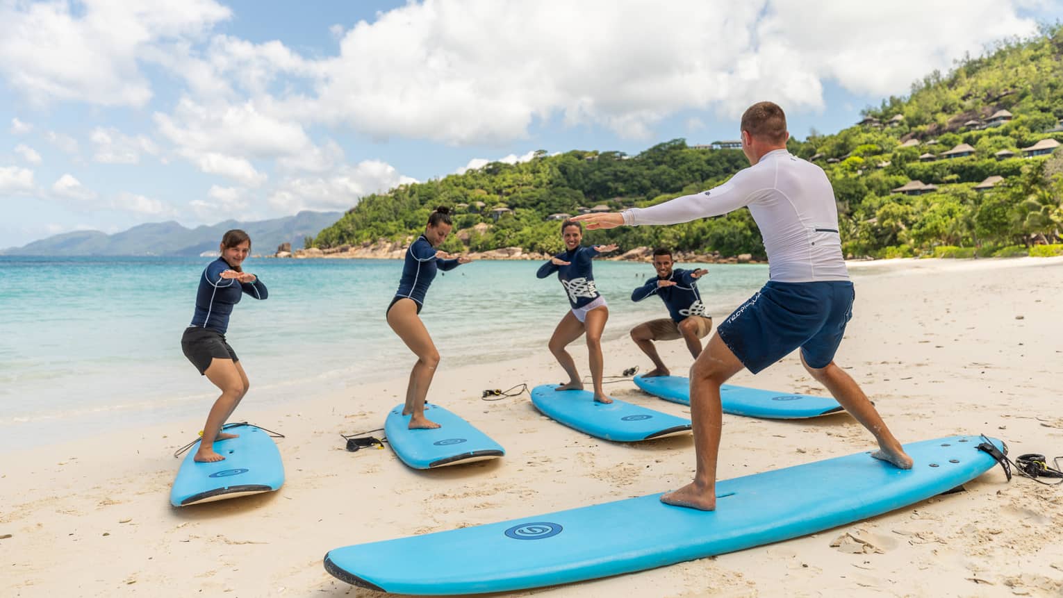 Surfing instructor balances on surfboard on white sand beach in front of row of surfers