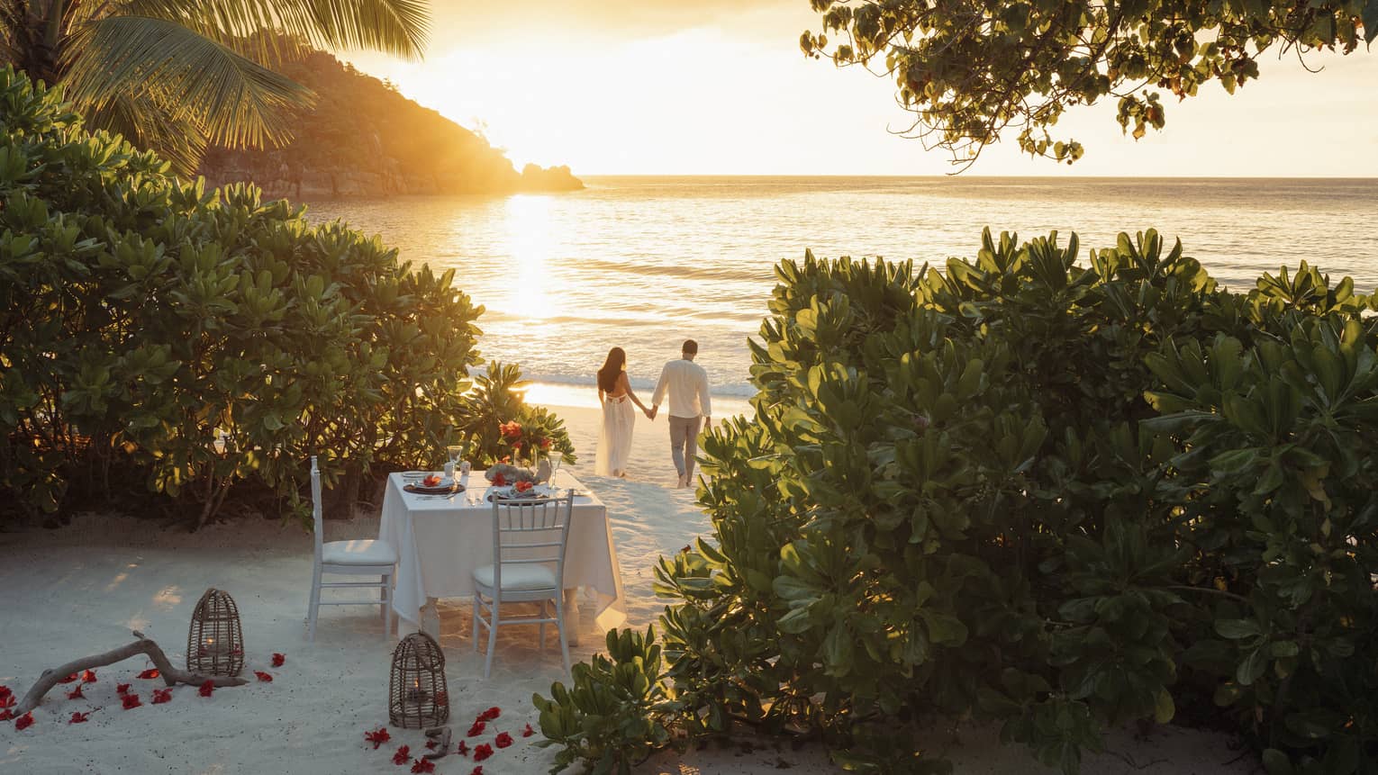 Framed by trees, a romantic dinner table set for two on the beach; a couple holding hands walks in the golden glow of sunset.