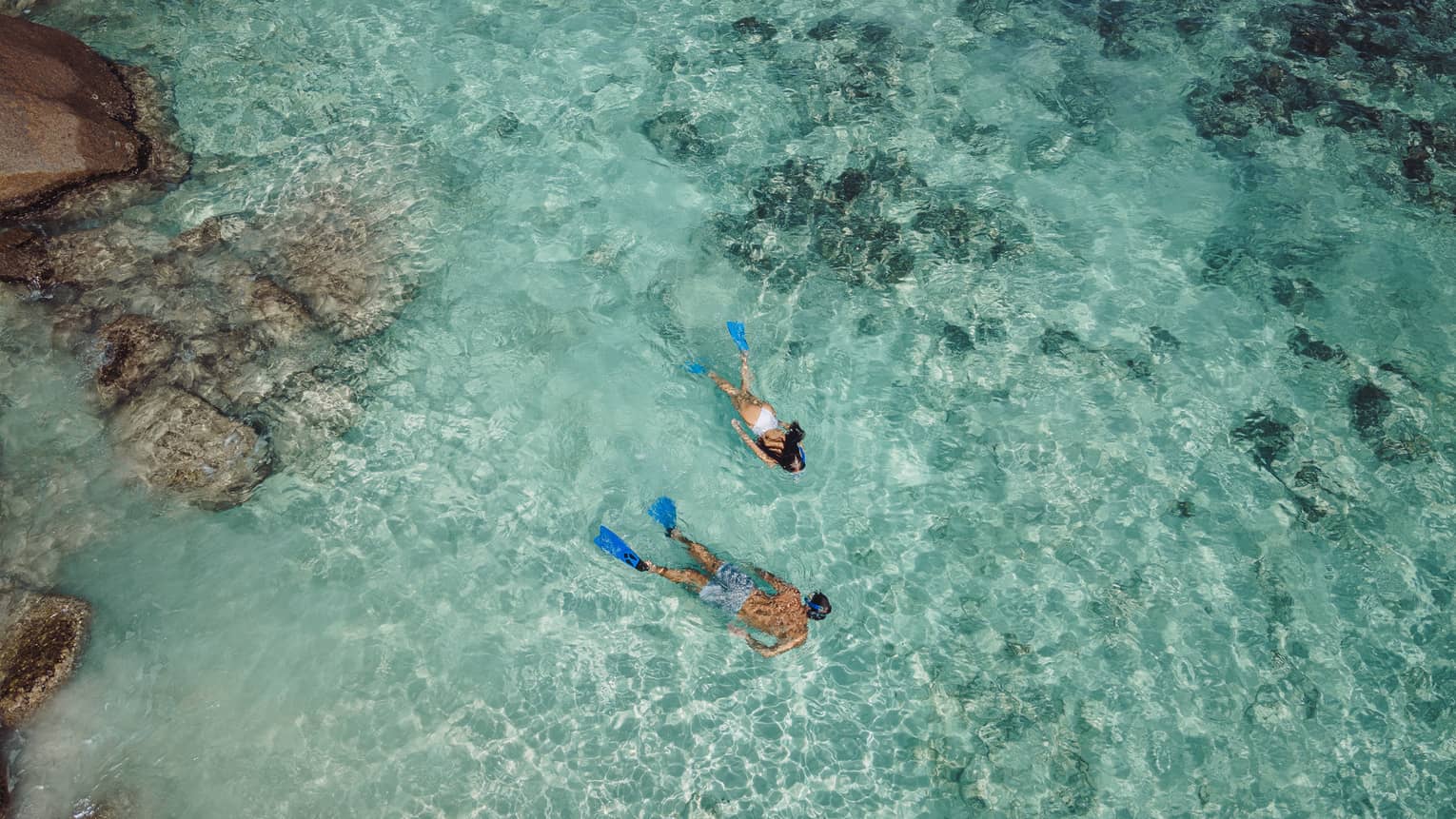 Aerial view of two snorkellers wearing blue flippers in an expanse of shallow, turquoise waters rippling over the reef.