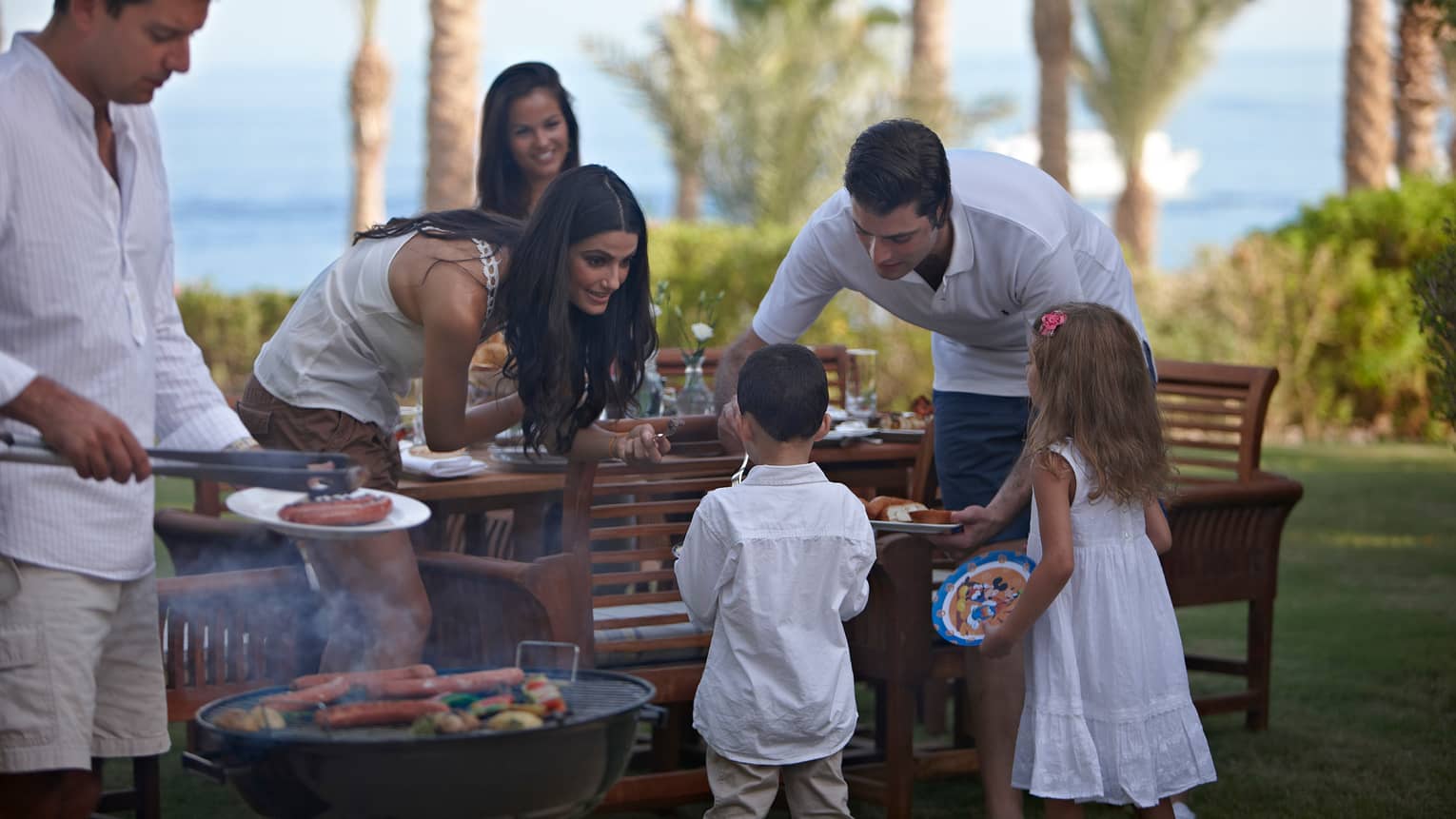 Man and woman dish out dinner for two children by patio table, man flips hot dogs on small barbecue