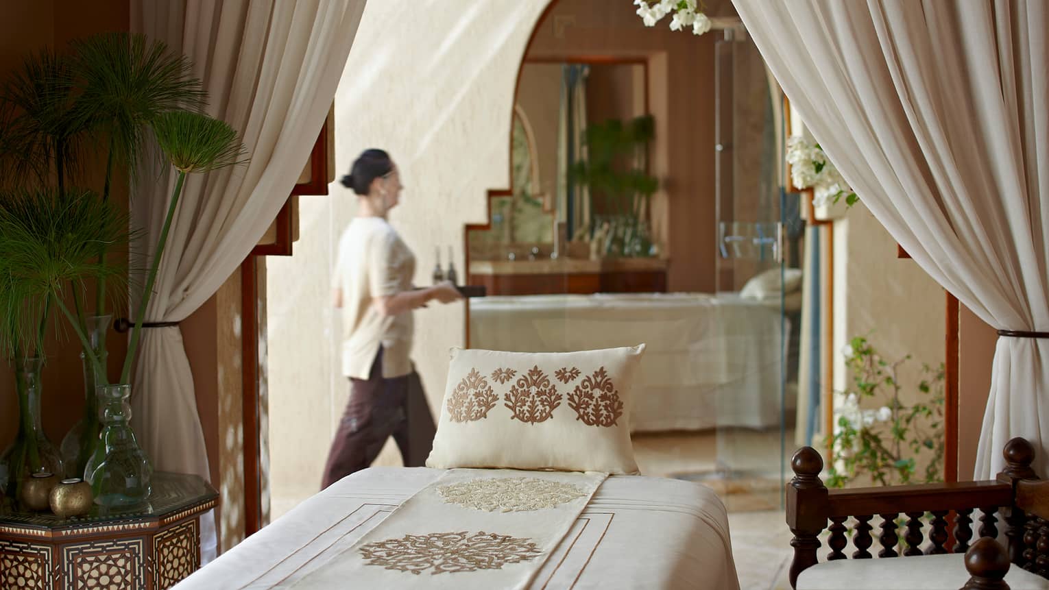Woman with tray walks behind massage bed with embroidered pillow in spa