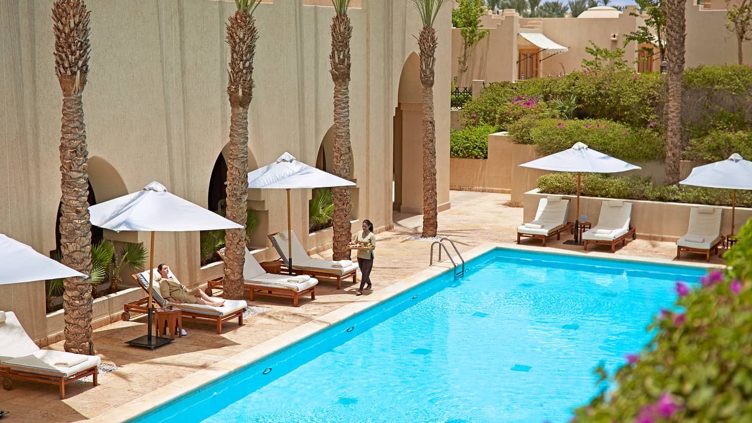 Server with tray walks along outdoor swimming pool deck, past woman lounging in white chair